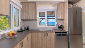A modern kitchen with wooden cabinets, a stainless steel refrigerator, a coffee machine, and a view of the sea through a window.