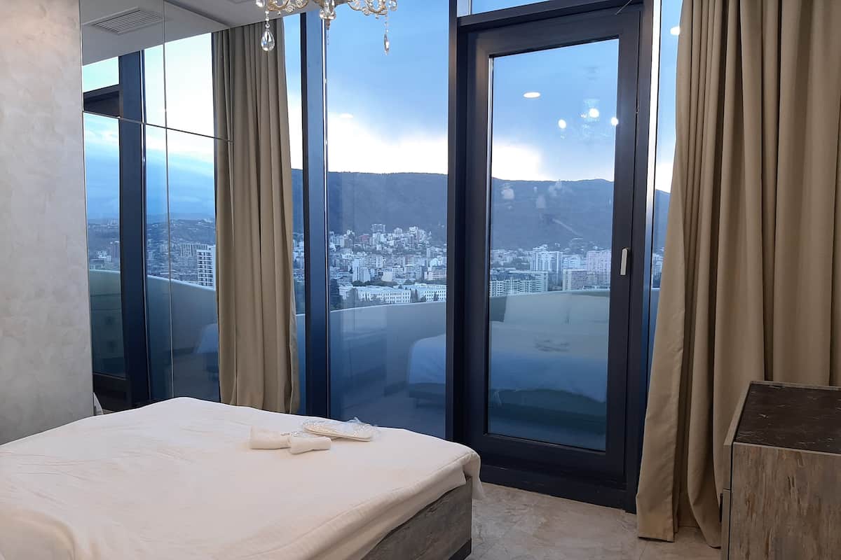 A bedroom with a large bed, a view of a cityscape through floor-to-ceiling windows, and a chandelier.