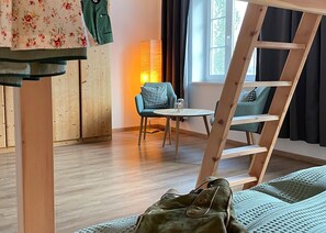 Un lit superposé avec un sac vert dessus, une petite table avec une chaise et une fenêtre avec des rideaux.