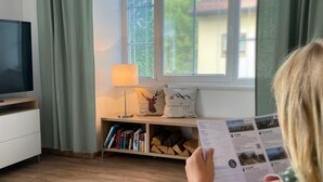 A room with a TV, books, a lamp, and a window with blinds. - Großzügige Ferienwohnung zum Entspannen und Wohlfühlen, 78 m², 4 Personen, Wlan (Siegsdorf)