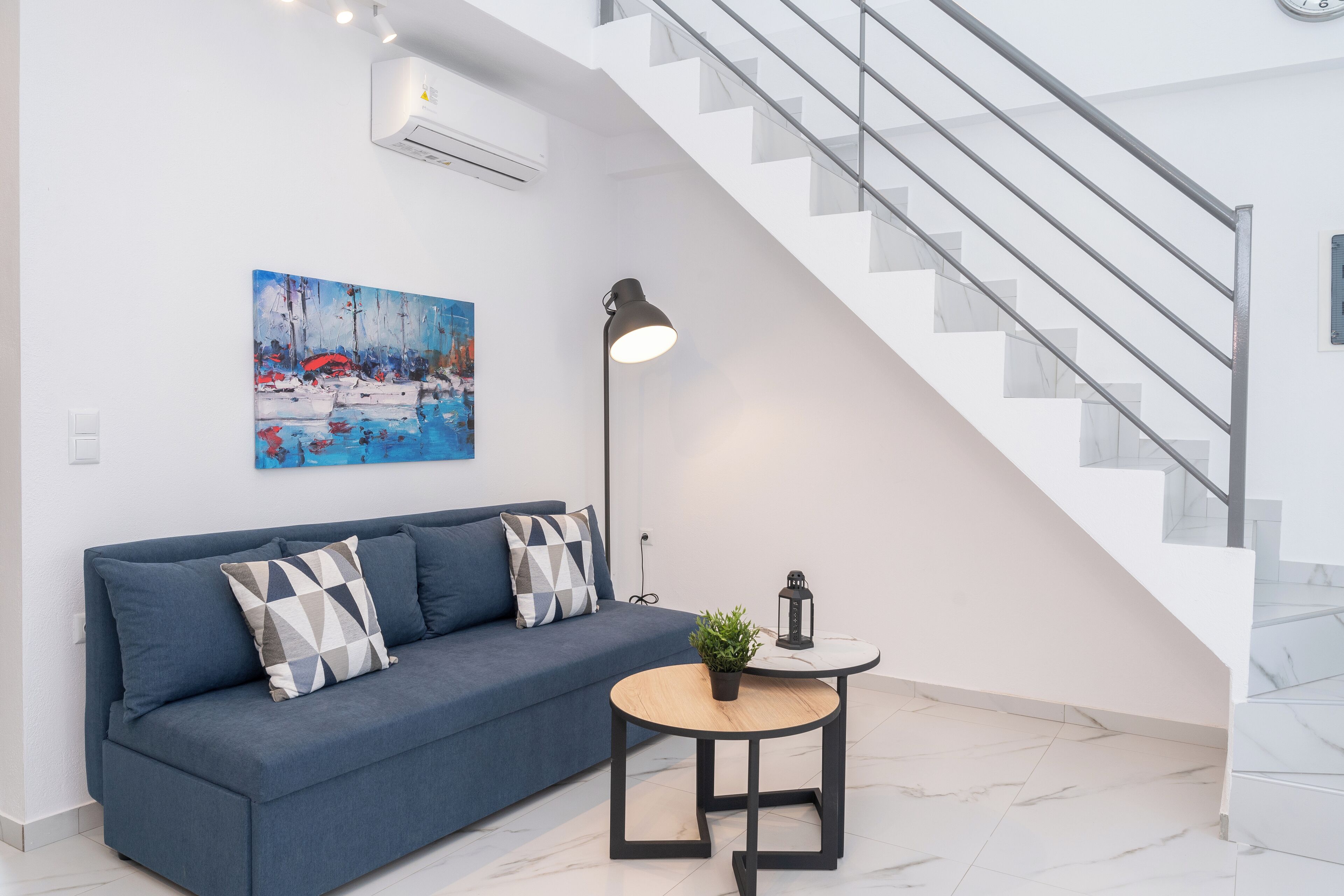 A modern living room with a blue sofa, a painting, a floor lamp, and a staircase.