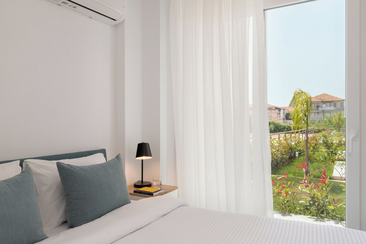 Een slaapkamer met een bed, kussens, een nachtkastje met een lamp en uitzicht op een tuin door een raam.