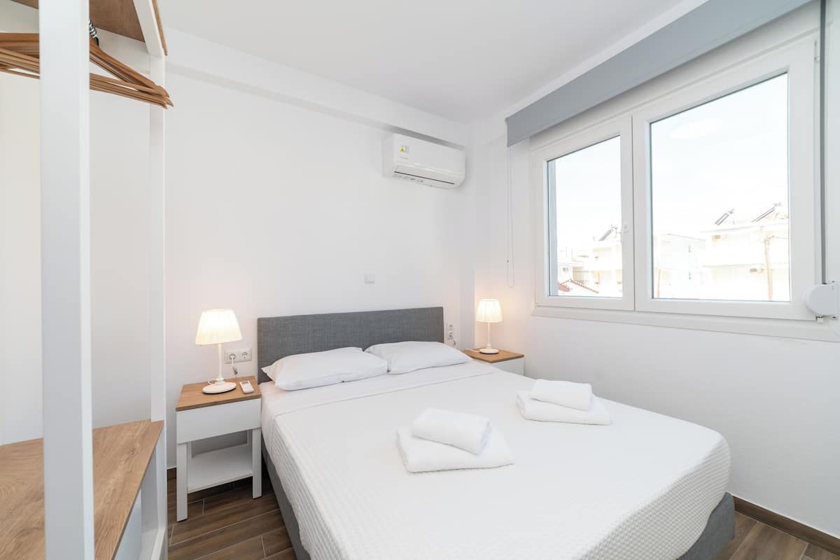 Een moderne hotelkamer met een groot bed, twee nachtkastjes met lampen, een grijs hoofdeinde en uitzicht op gebouwen door het raam.