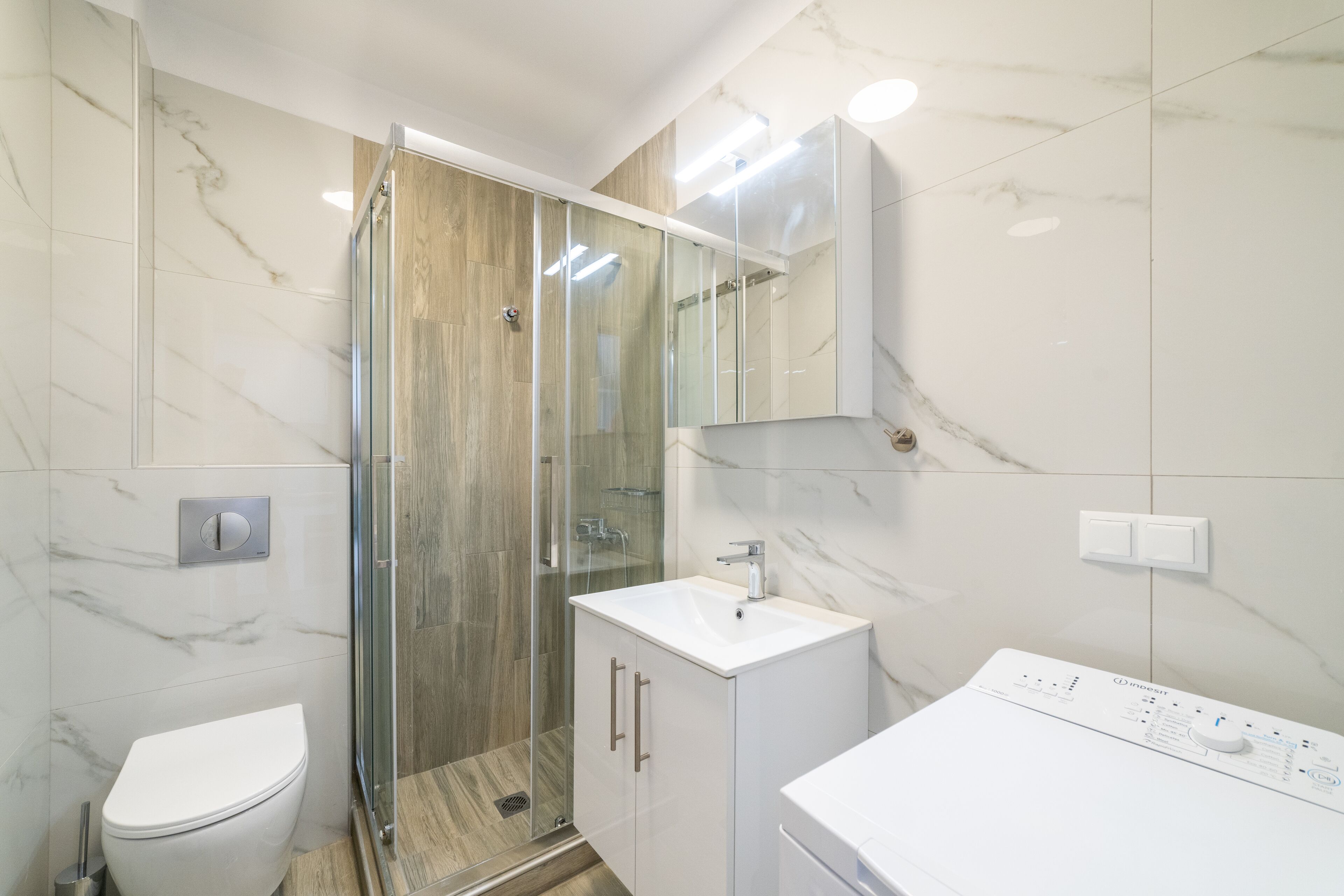 Casa de banho moderna com chuveiro envidraçado, sanita branca e lavatório branco com painel de controlo.