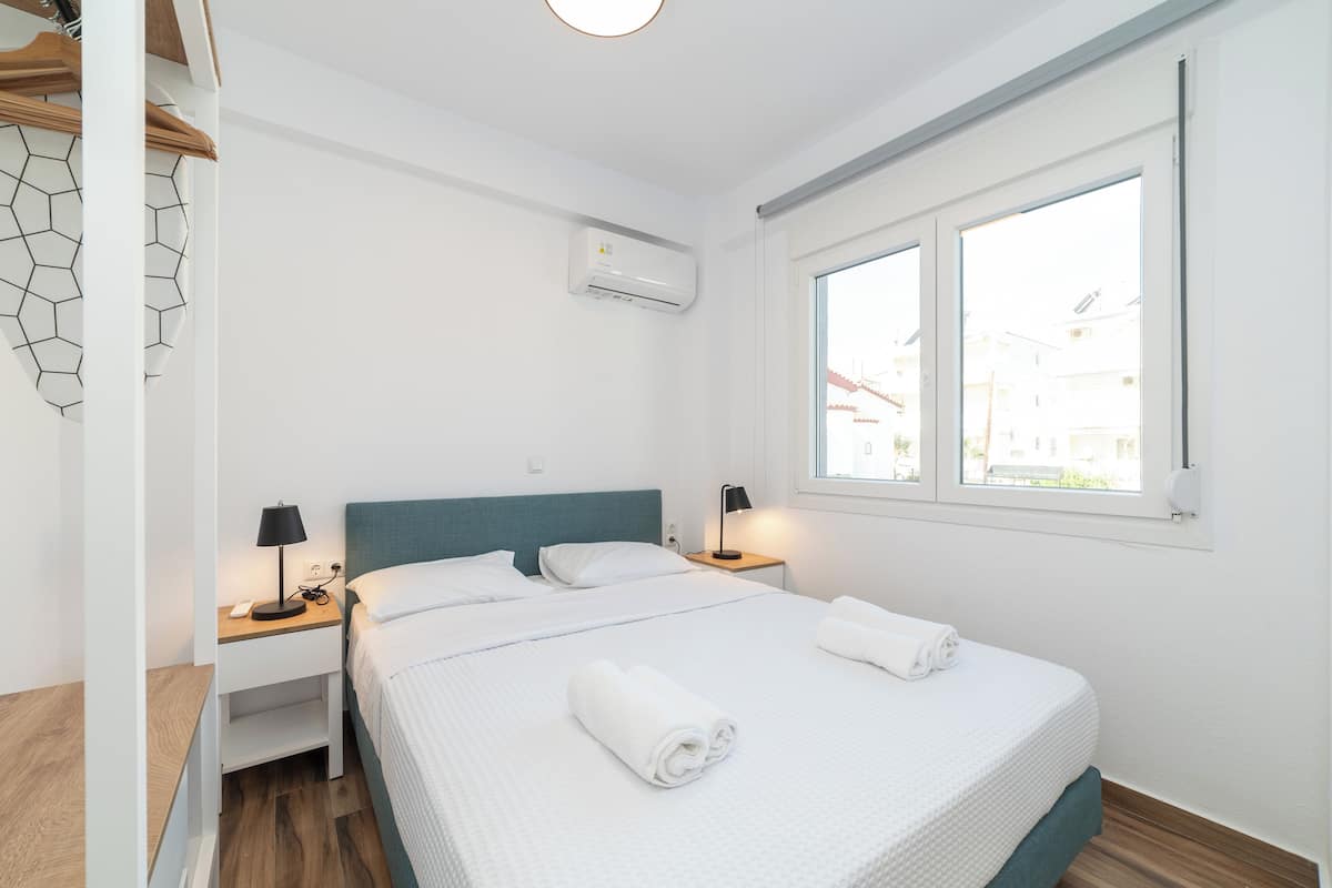 Een moderne hotelkamer met een groot bed, twee nachtkastjes, een wandairconditioner en een raam met uitzicht op gebouwen.