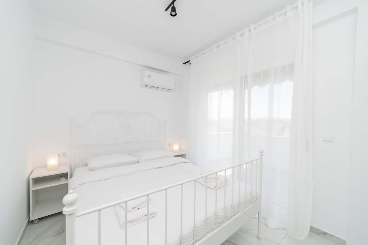 Een witte slaapkamer met een enkel bed, een nachtkastje en een airconditioner aan de muur.