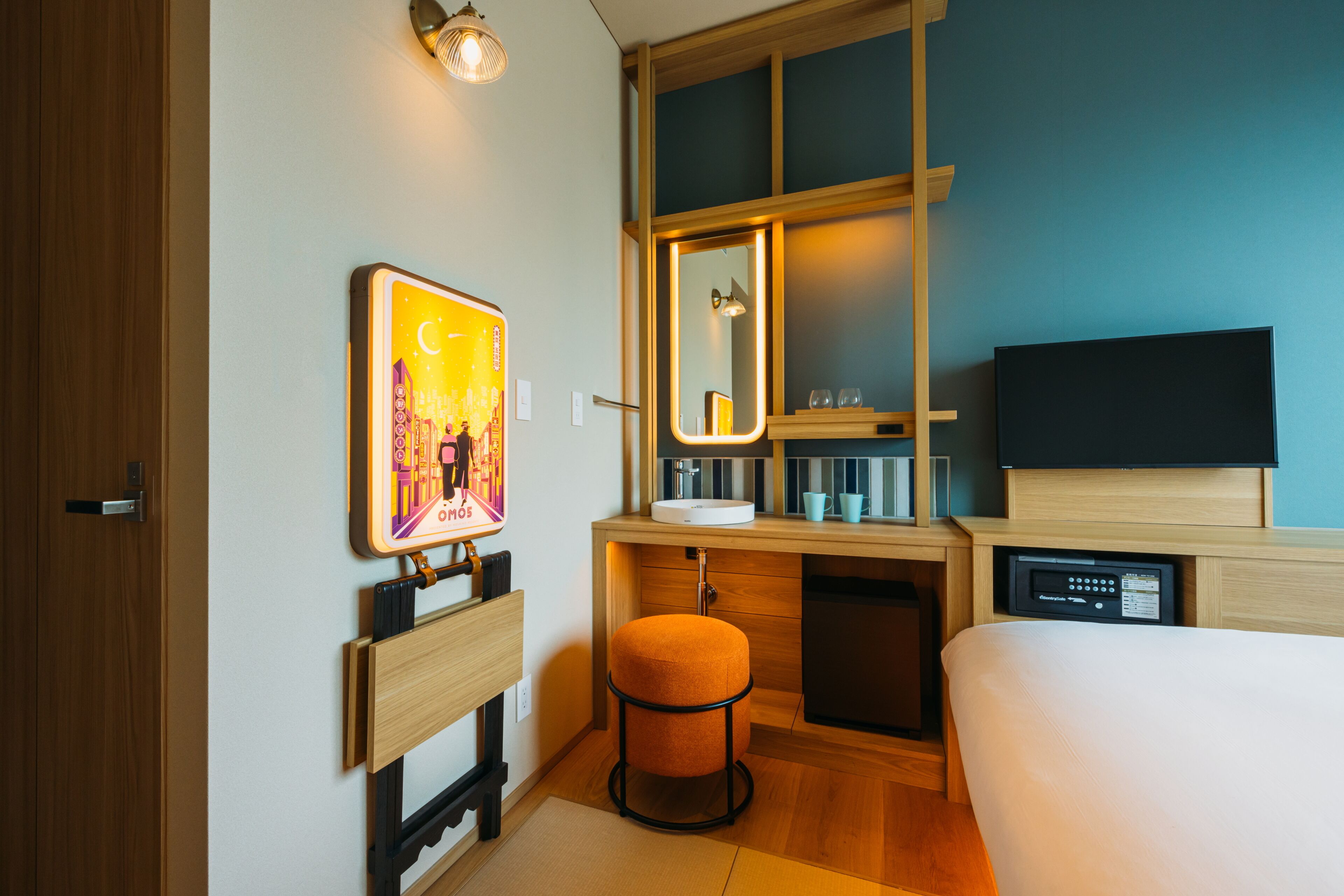 A hotel room with a wooden desk, a small stool, a bed, a television, a mirror, and a wall-mounted artwork.