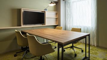 Une salle de conférence avec une table en bois, quatre chaises, un téléviseur fixé au mur et une fenêtre ornée de rideaux légers.