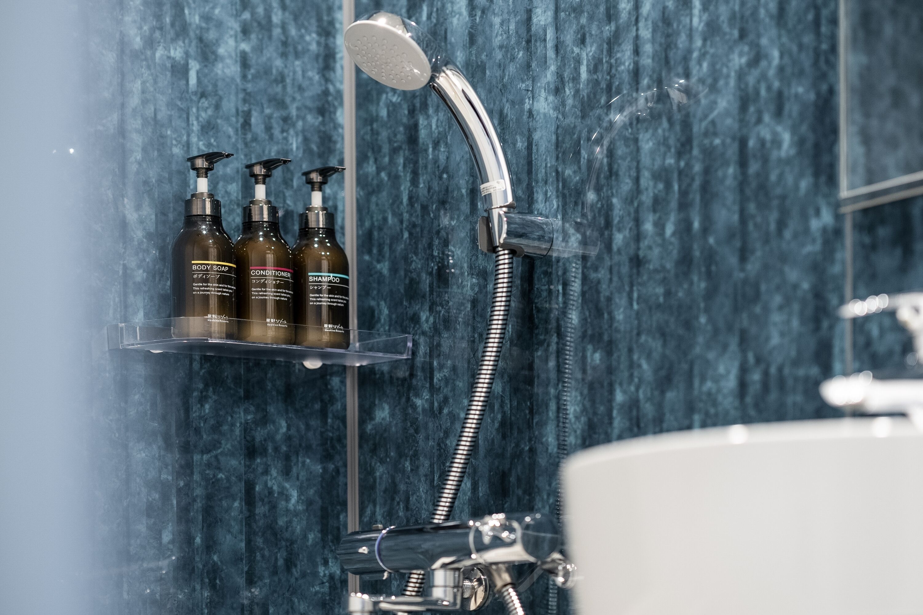 Casa de banho com chuveiro, lavatório e três frascos de shampoo e condicionador numa prateleira.