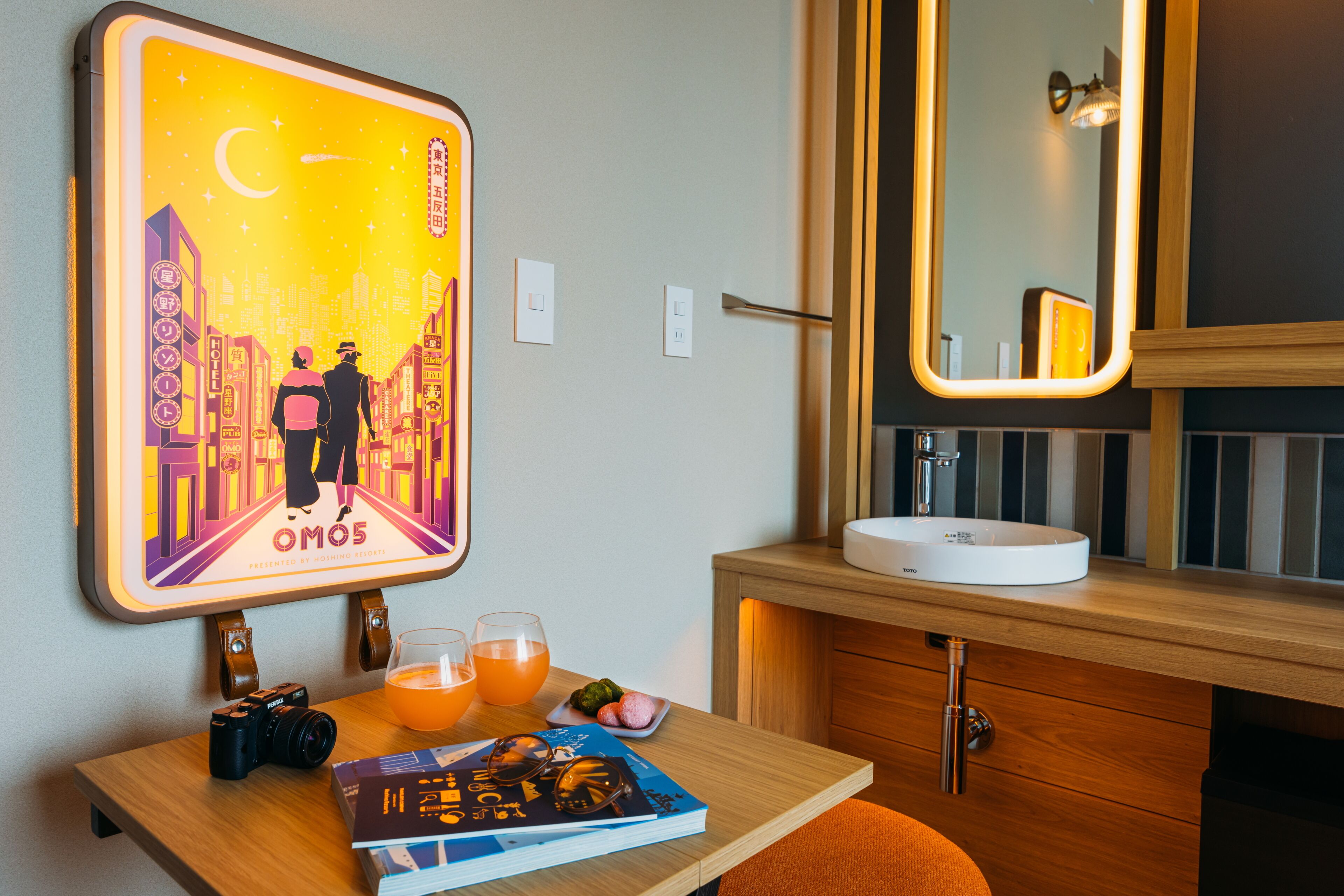 A hotel room with a wooden vanity, a mirror, a table with a camera, glasses, a book, and a poster on the wall.