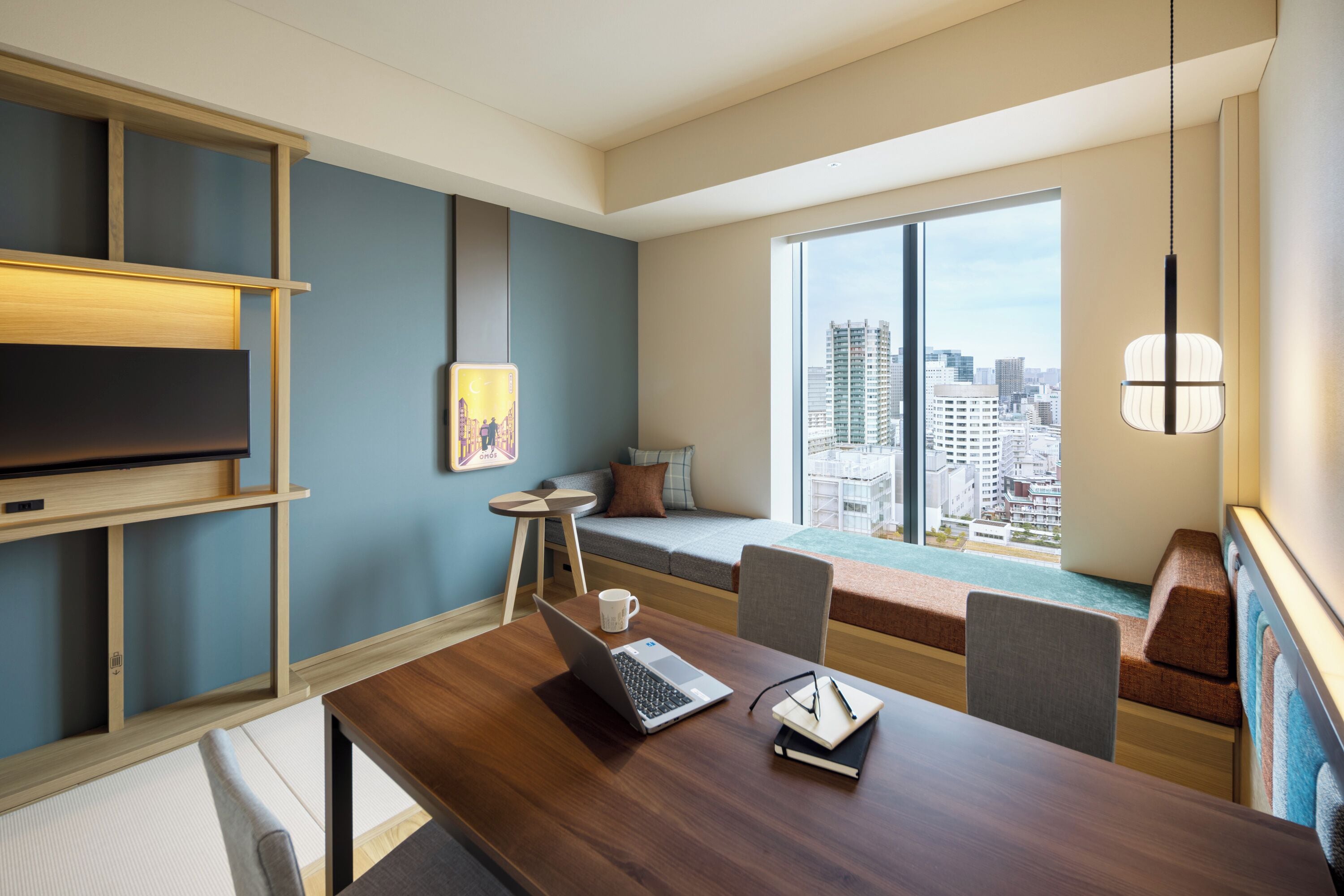 A modern hotel room with a wooden desk, a laptop, a notebook, and a cup. There is a large window with a city view.