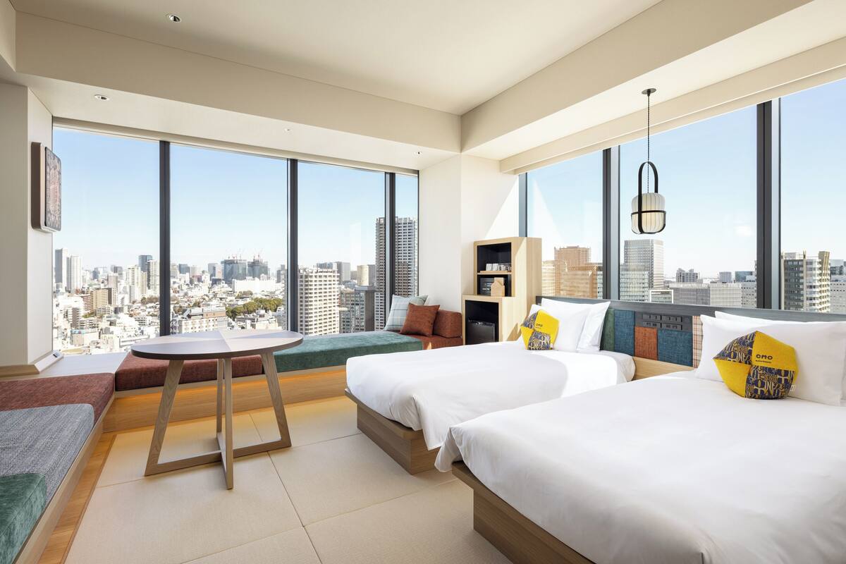Une chambre d’hôtel moderne avec deux lits, une petite table, un canapé et une vue sur le paysage urbain.