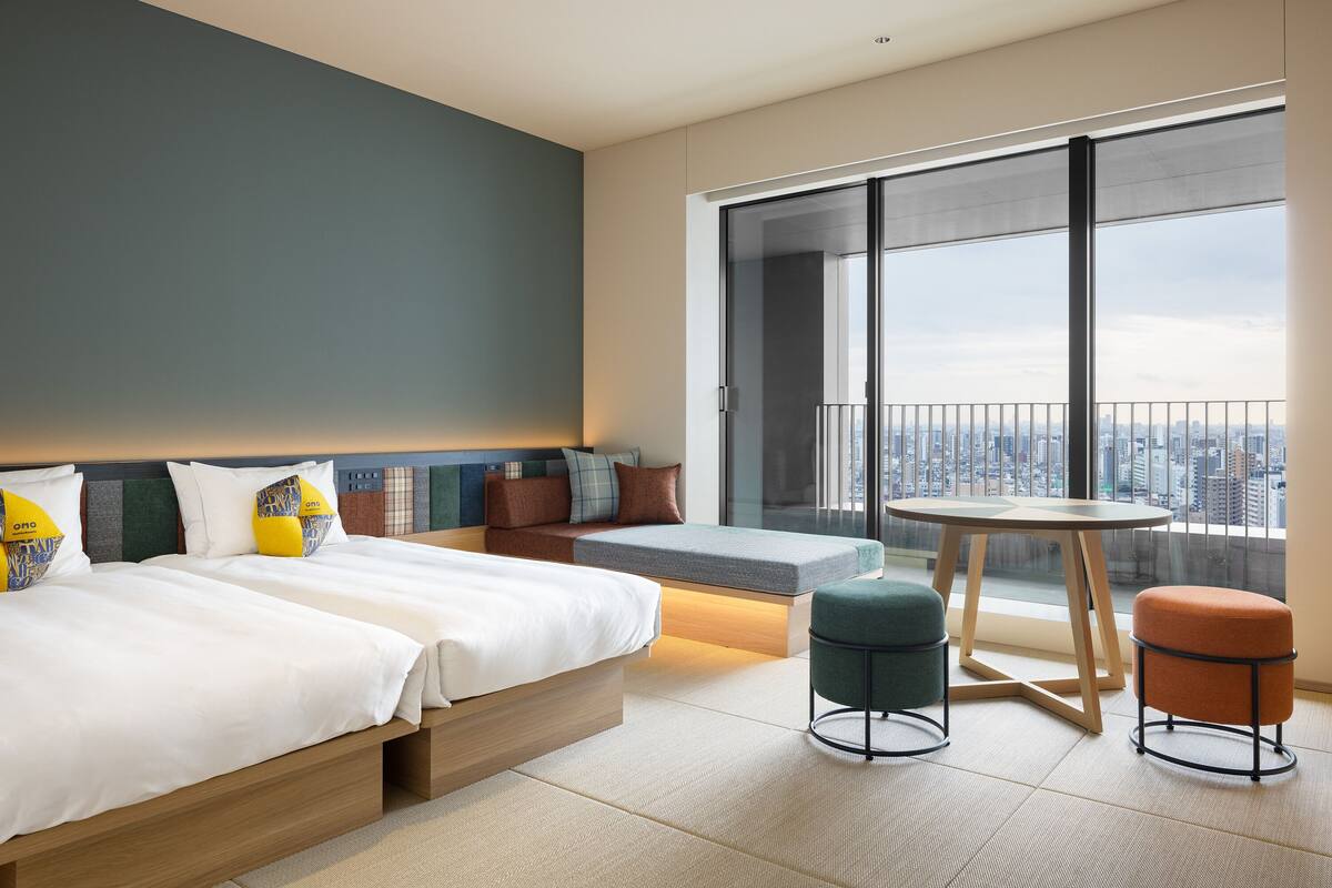 Une chambre d’hôtel moderne dotée d’une grande fenêtre donnant sur la ville, d’un lit avec du linge de lit blanc, d’une petite table et de deux poufs.