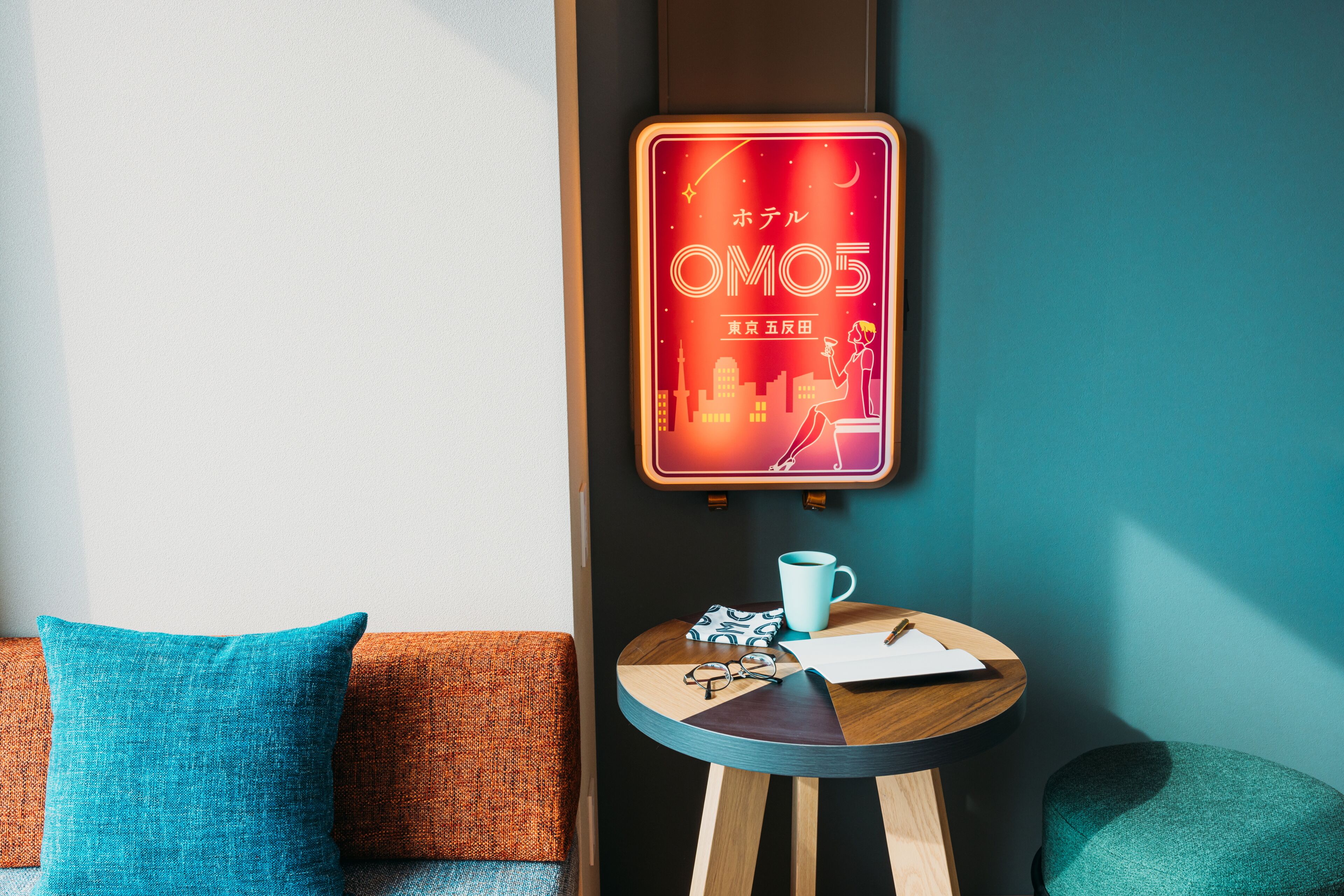 A cozy room with a teal sofa, a small wooden table with a cup, glasses, and a notepad. A neon sign with Japanese characters is on the wall.