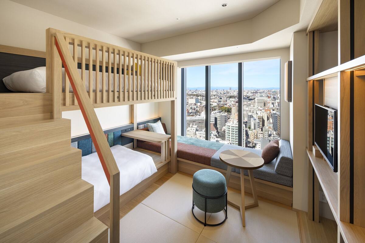 Une chambre d’hôtel moderne avec un lit superposé en bois, un petit coin salon avec une table et une grande fenêtre donnant sur la ville.