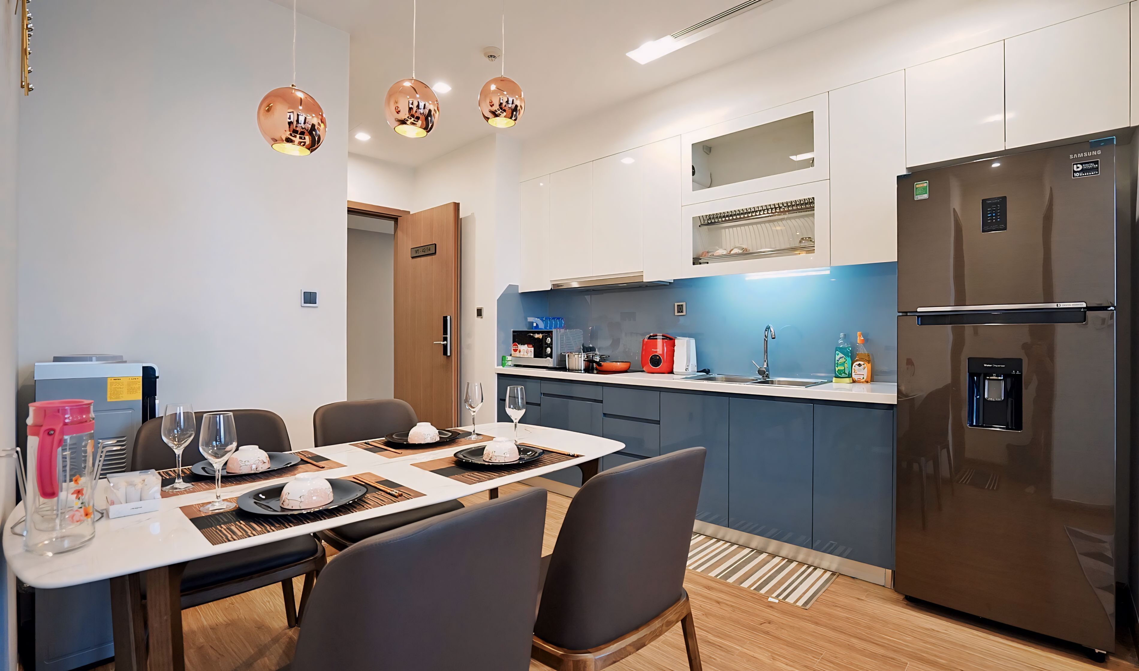 A modern kitchen with a stainless steel refrigerator, a microwave, and a coffee maker. There is a dining table with chairs and a small island with a sink.