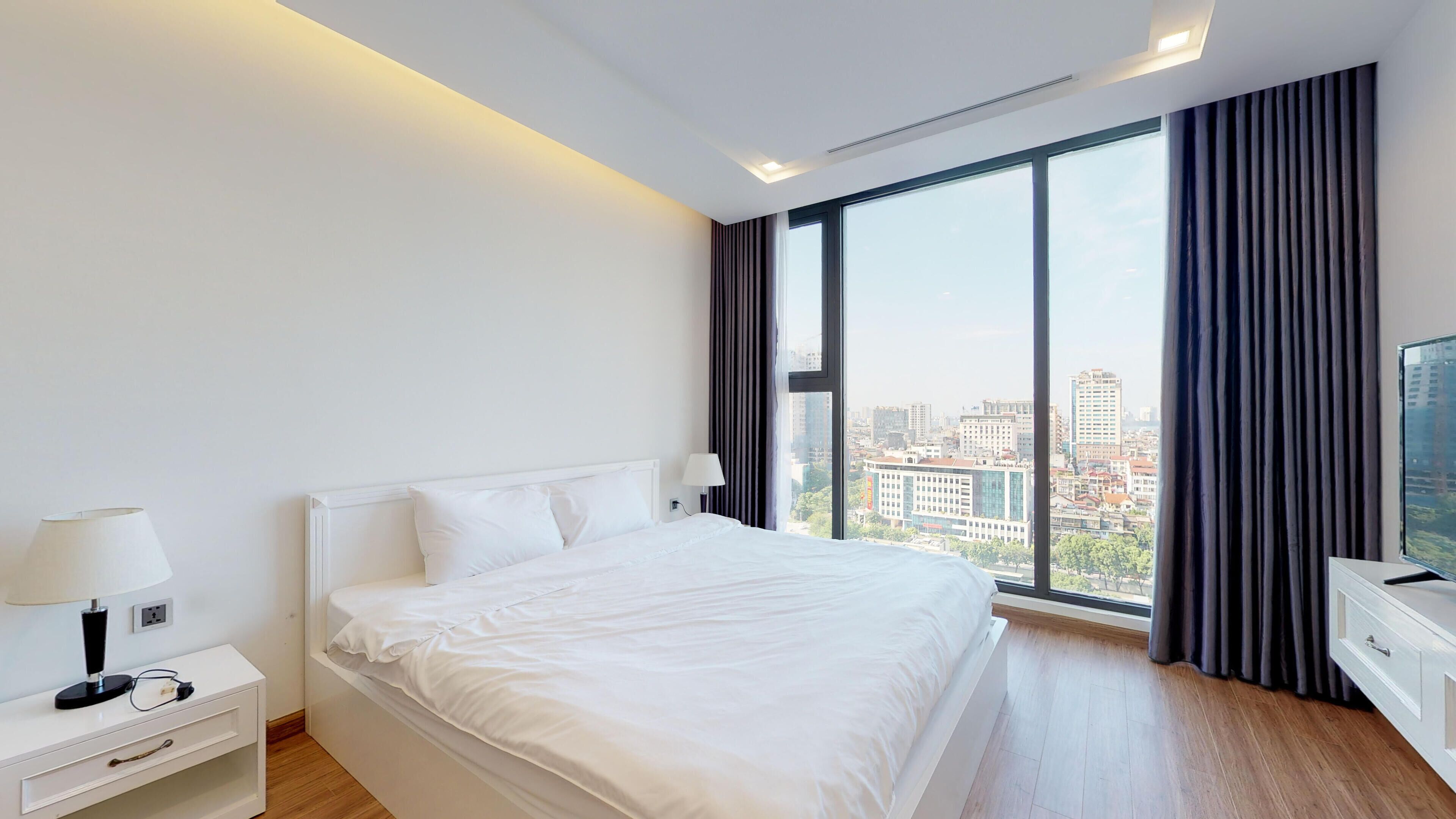 A modern hotel room with a large bed, a bedside table with a lamp, a dresser, and a view of the city through large windows.