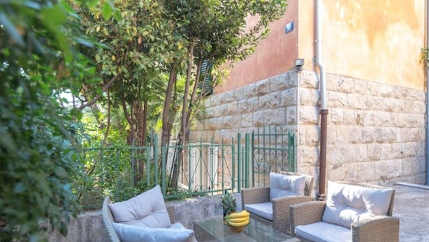 Un espacio de estar al aire libre con mobiliario de mimbre y cojines, una mesa de vidrio con frutero, y un muro de piedra.