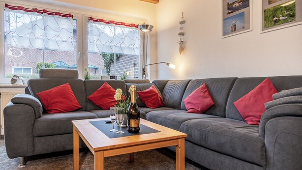 A living room with a grey sofa, red cushions, a wooden coffee table with a bottle and glasses, and a view of a brick building through the windows.