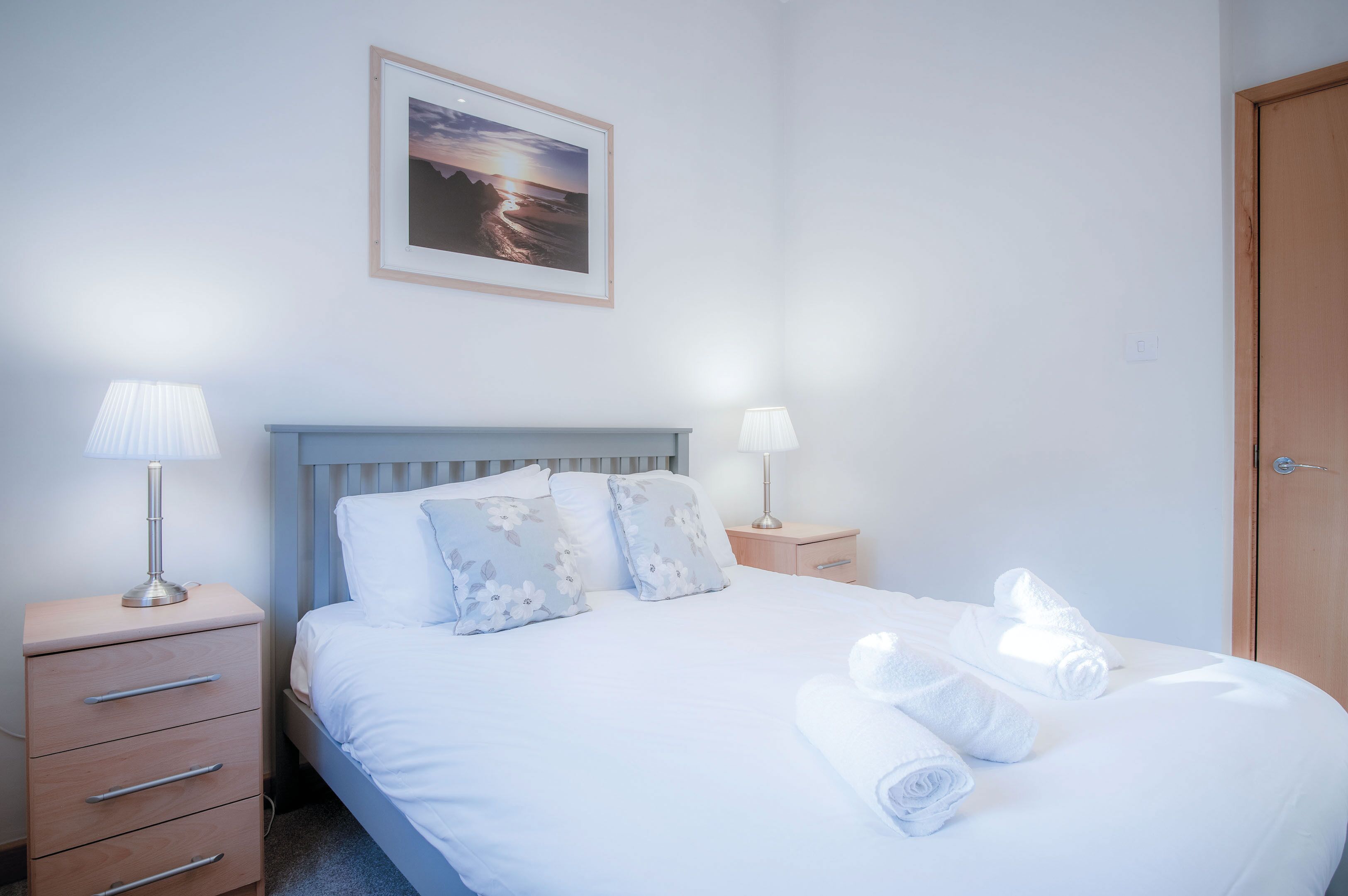 Ein ordentlich bezogenes Bett mit floralen Kissen, einer Nachttischlampe und aufgerollten Handtüchern auf einem Holznachttisch.