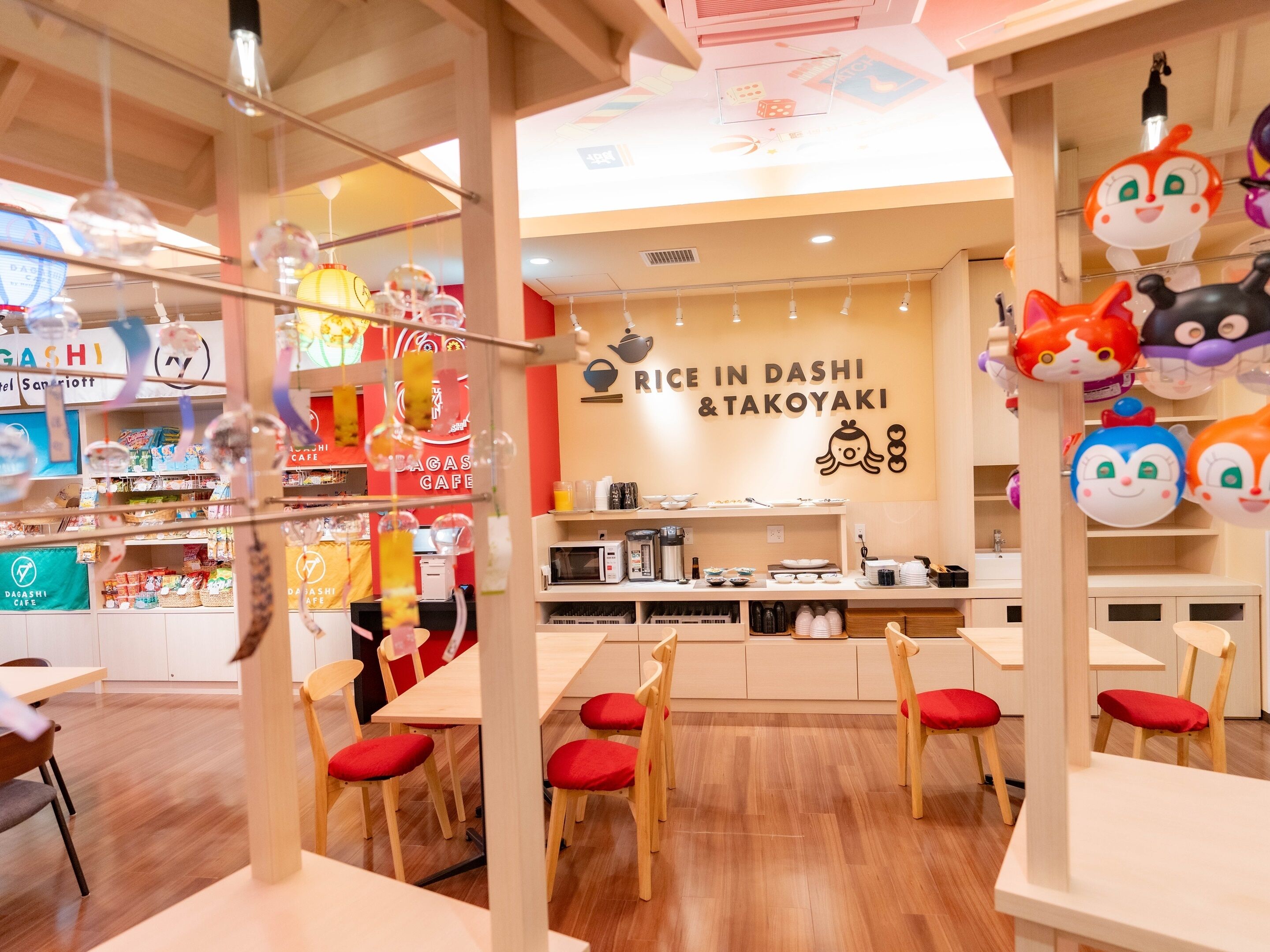 Interior de una cafetería con mesas y sillas de madera, un mostrador con electrodomésticos de cocina y una pared con un letrero que dice 'Rice in Dashi & Takoyaki'.