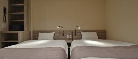 A hotel room with two beds, a wooden shelf unit, and a wall-mounted telephone.