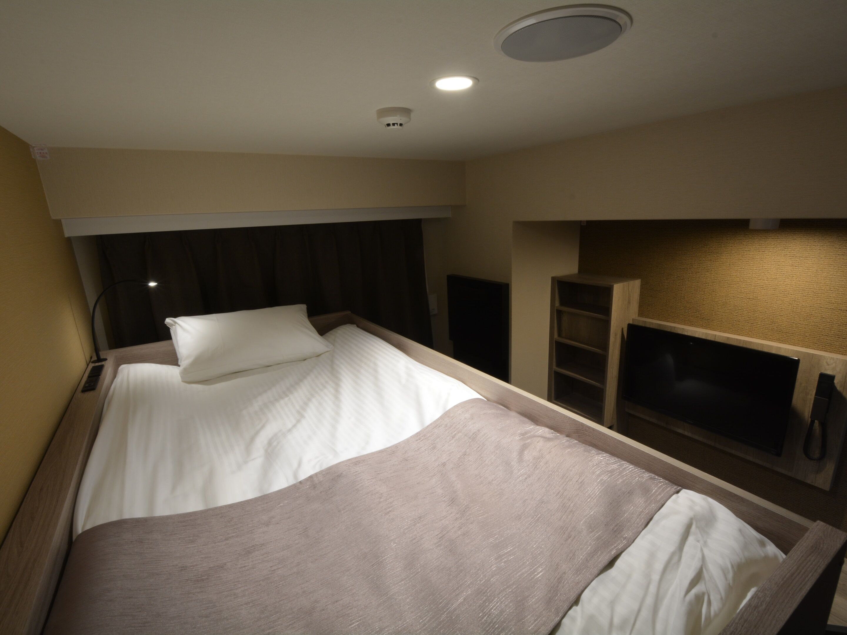 A compact hotel room with a single bed, a bedside table, a lamp, and a built-in TV shelf.