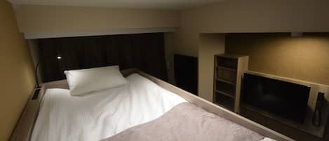 A compact hotel room with a single bed, a bedside table, a lamp, and a built-in TV shelf.