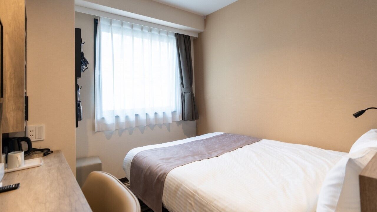 A hotel room with a bed, a desk with a chair, a window with curtains, and a wall-mounted lamp.