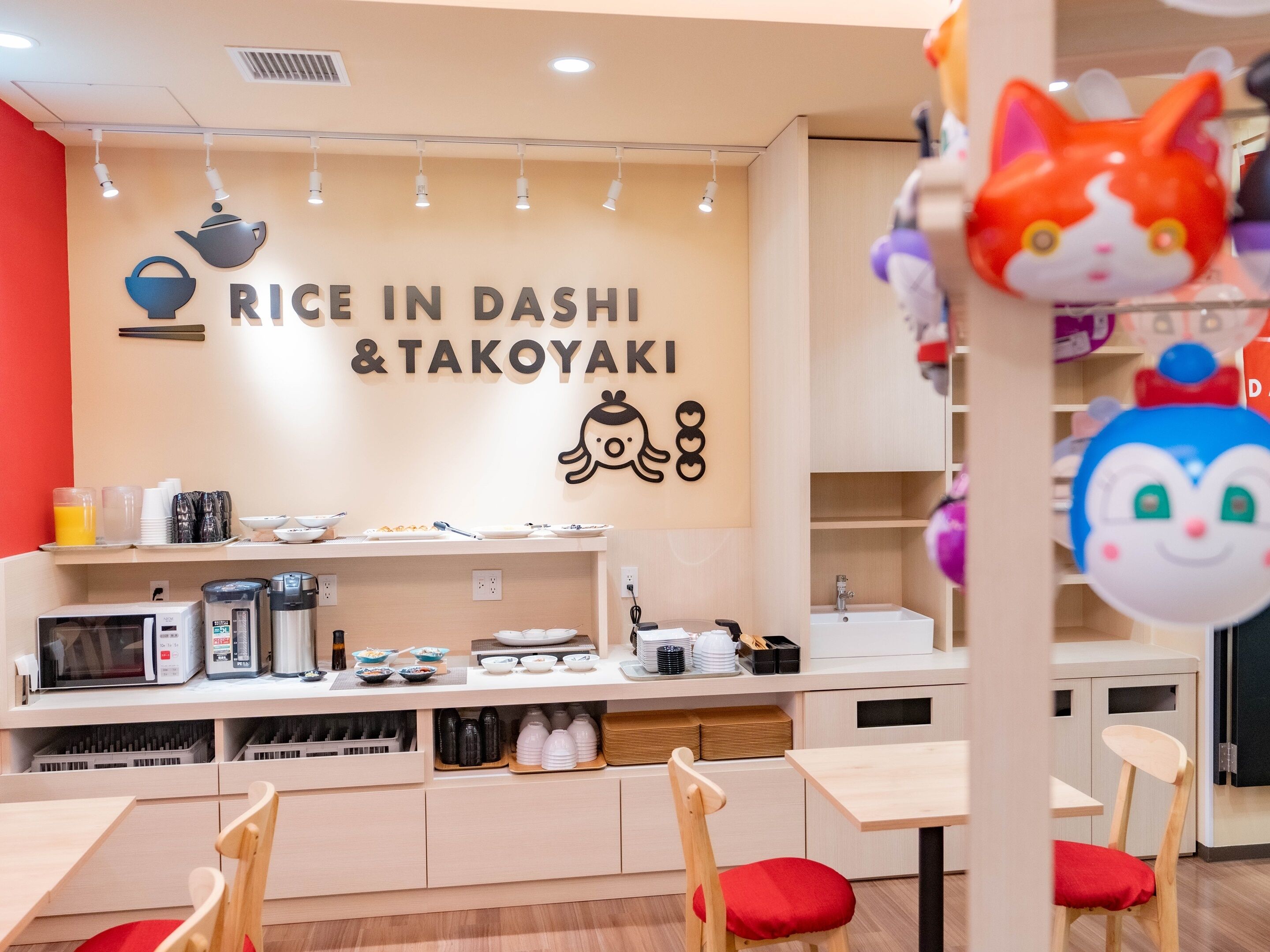 A modern dining area with a wall sign reading "RICE IN DASHI & TAKOYAKI," a microwave, and various dishes on a counter.