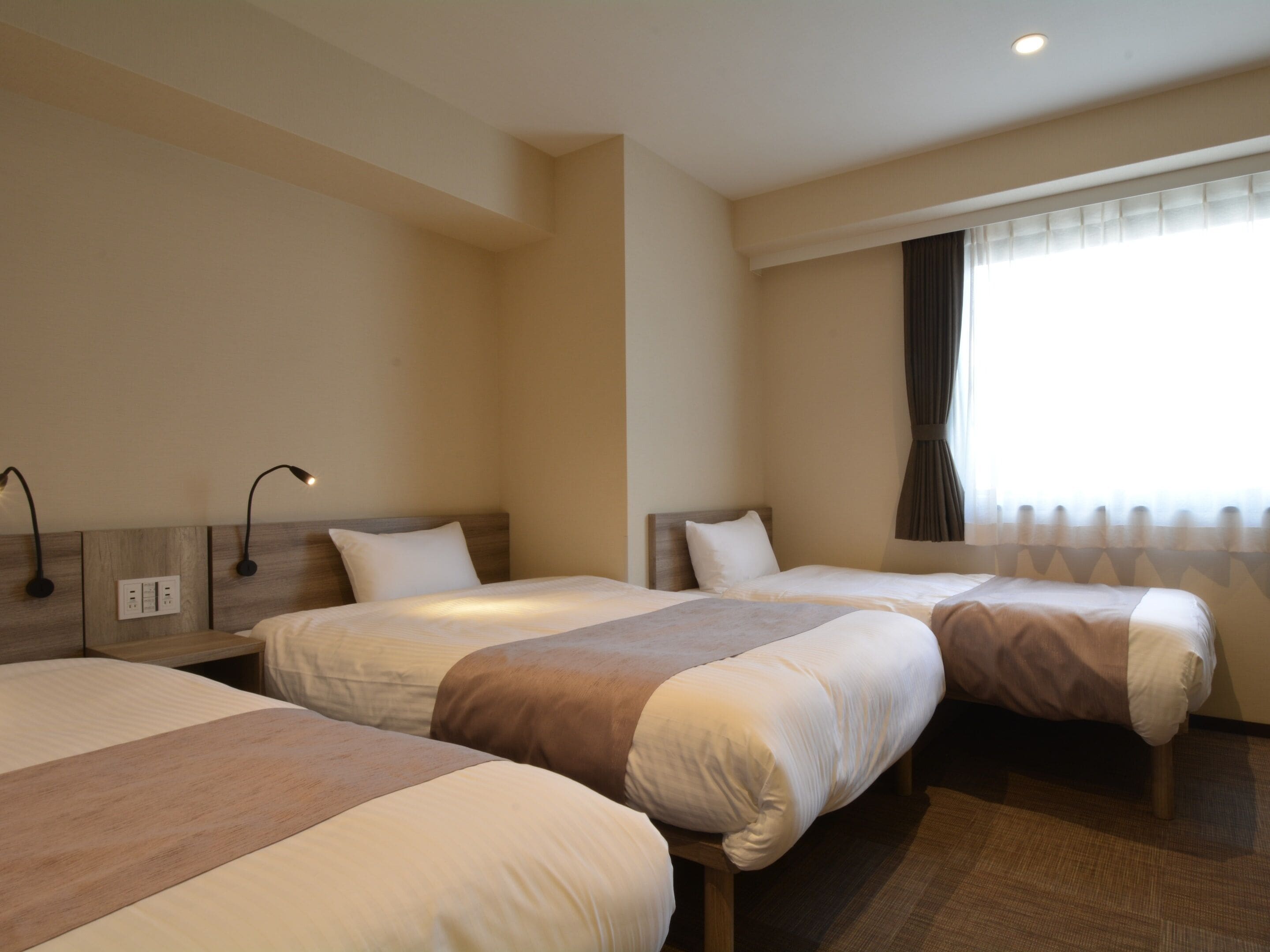 A hotel room with two beds, a window with curtains, and a wall-mounted lamp.