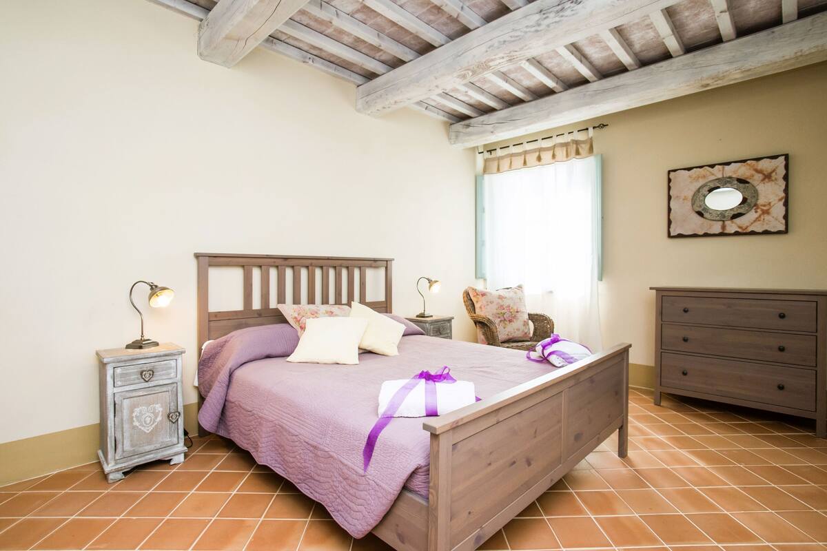 A bedroom with a wooden bed, a nightstand with a lamp, a dresser, and a window with curtains.