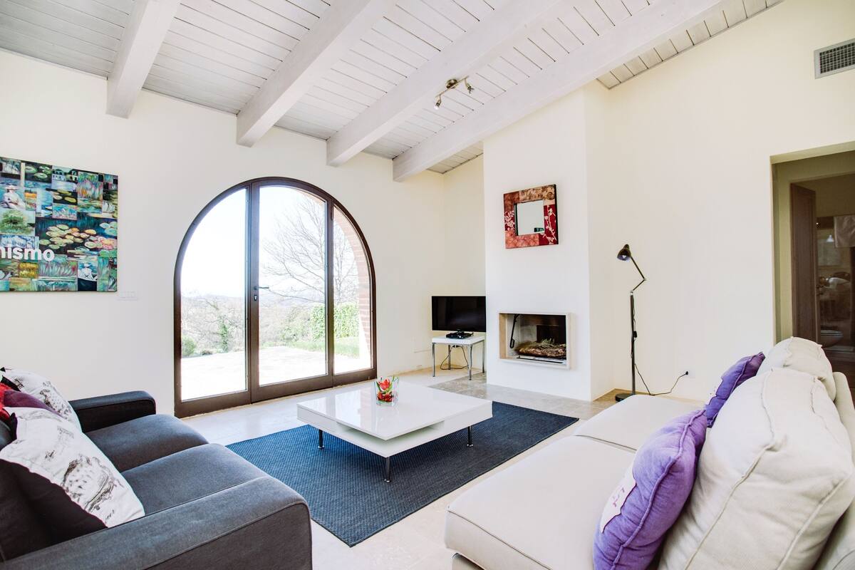 A modern living room with a large arched window, a flat-screen TV, a fireplace, and a cozy seating area.