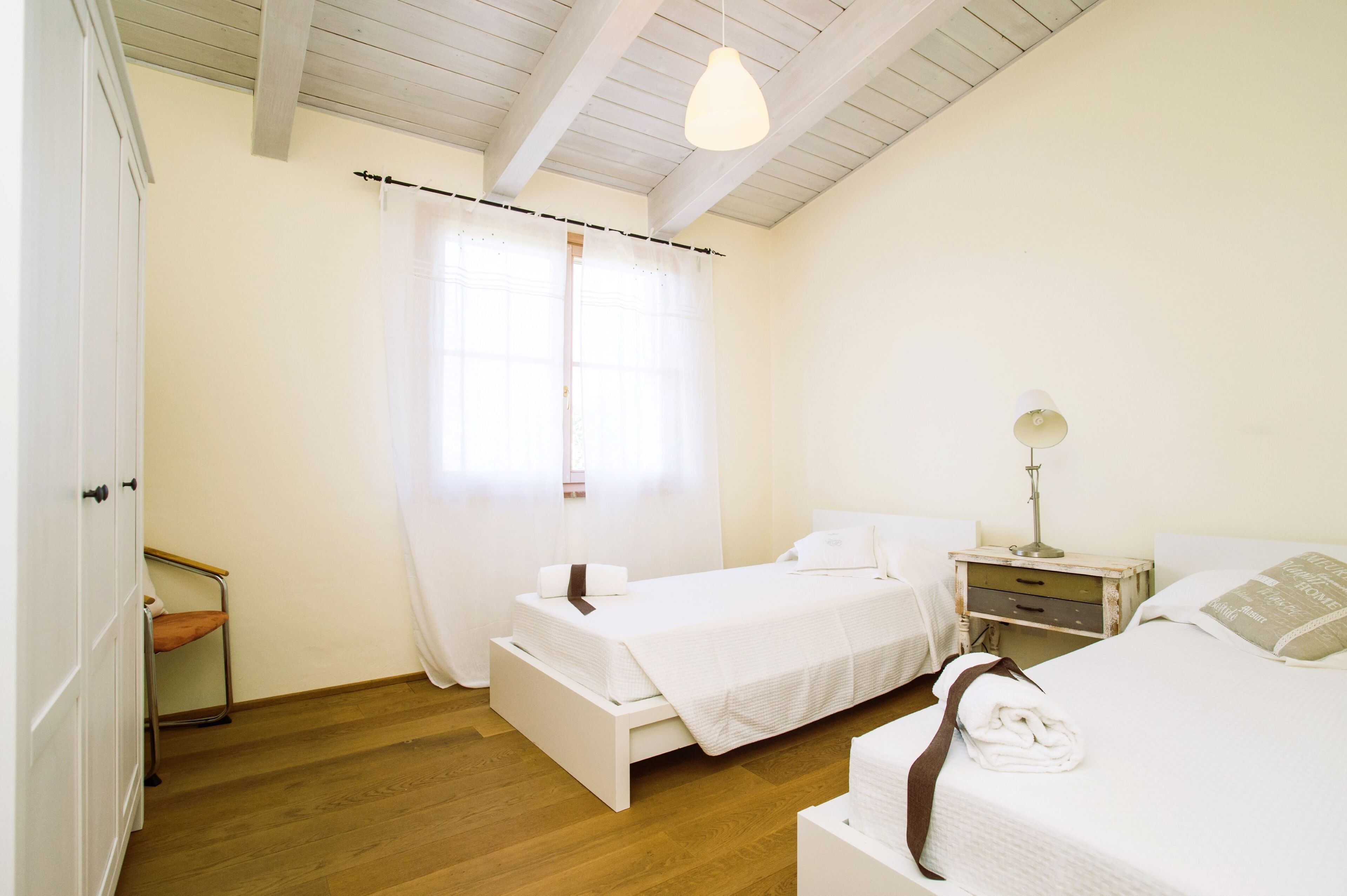 A bedroom with two beds, a wooden floor, a white wardrobe, and a bedside table with a lamp.