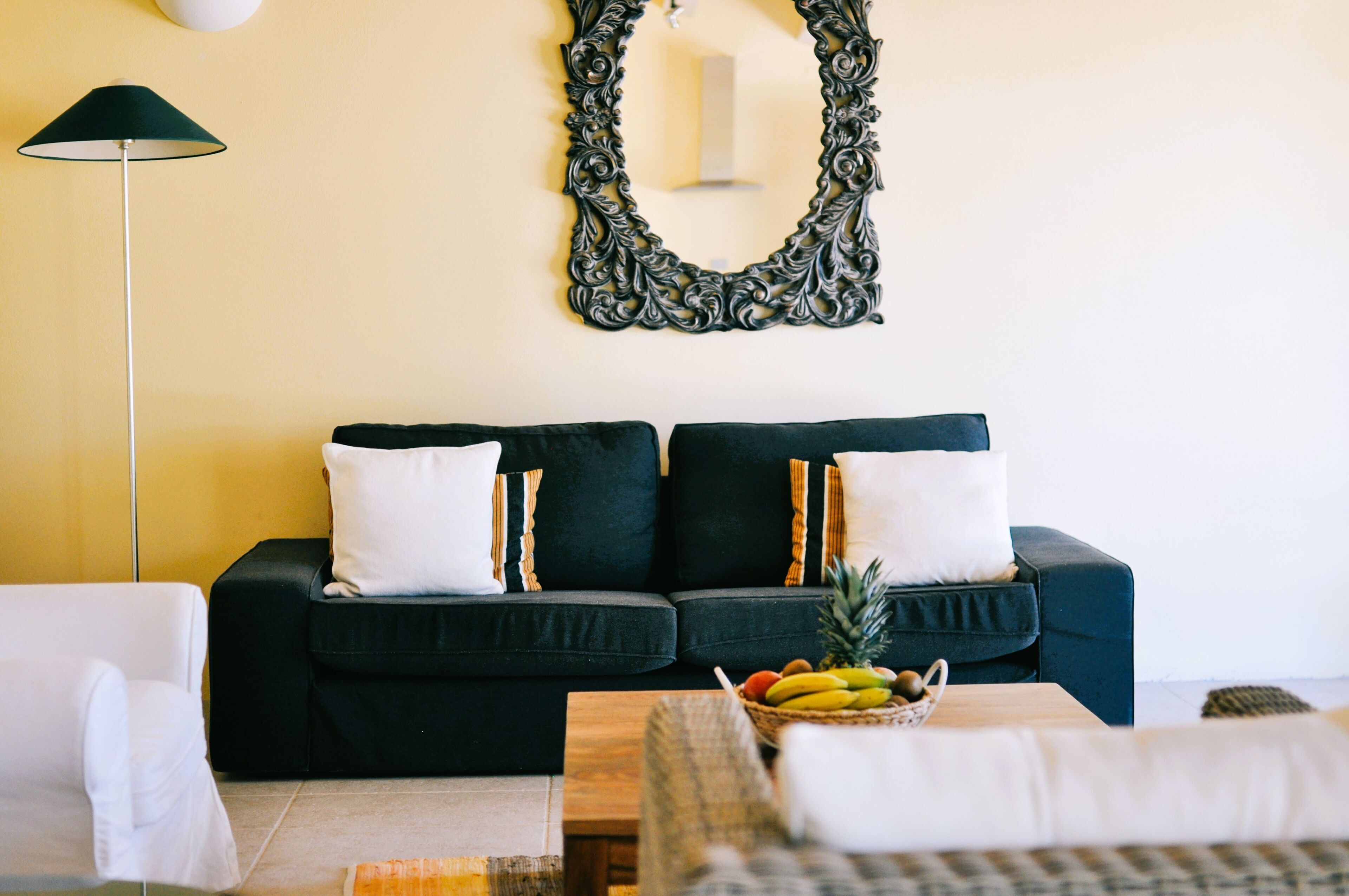 A living room with a dark green sofa, white cushions, a floor lamp, and a fruit bowl on a wooden table.