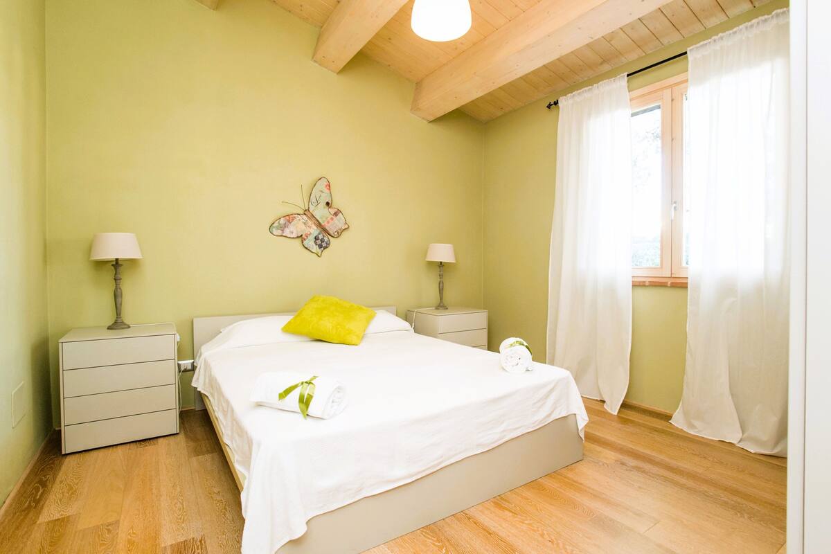 A bedroom with a bed, two bedside tables, a nightstand, a wall-mounted lamp, a window with curtains, and a butterfly decoration.