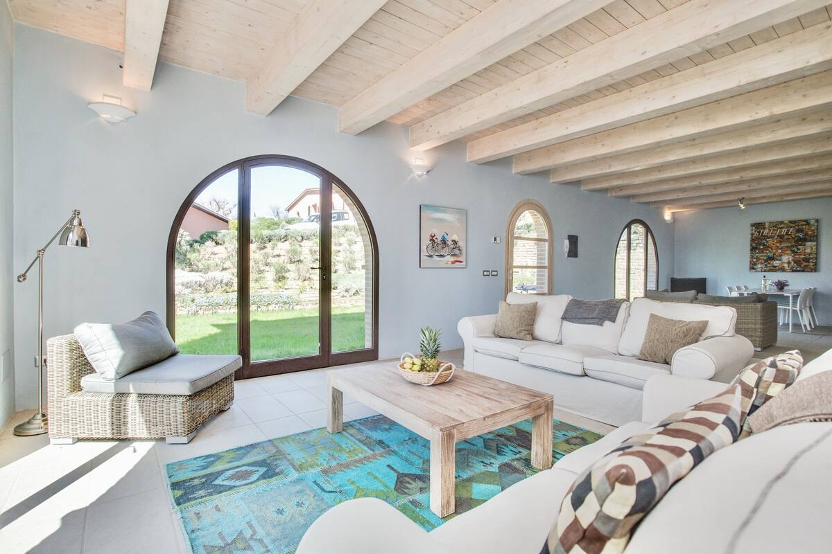 A spacious living room with a large window, a white sofa, a wicker chair, and a wooden coffee table.
