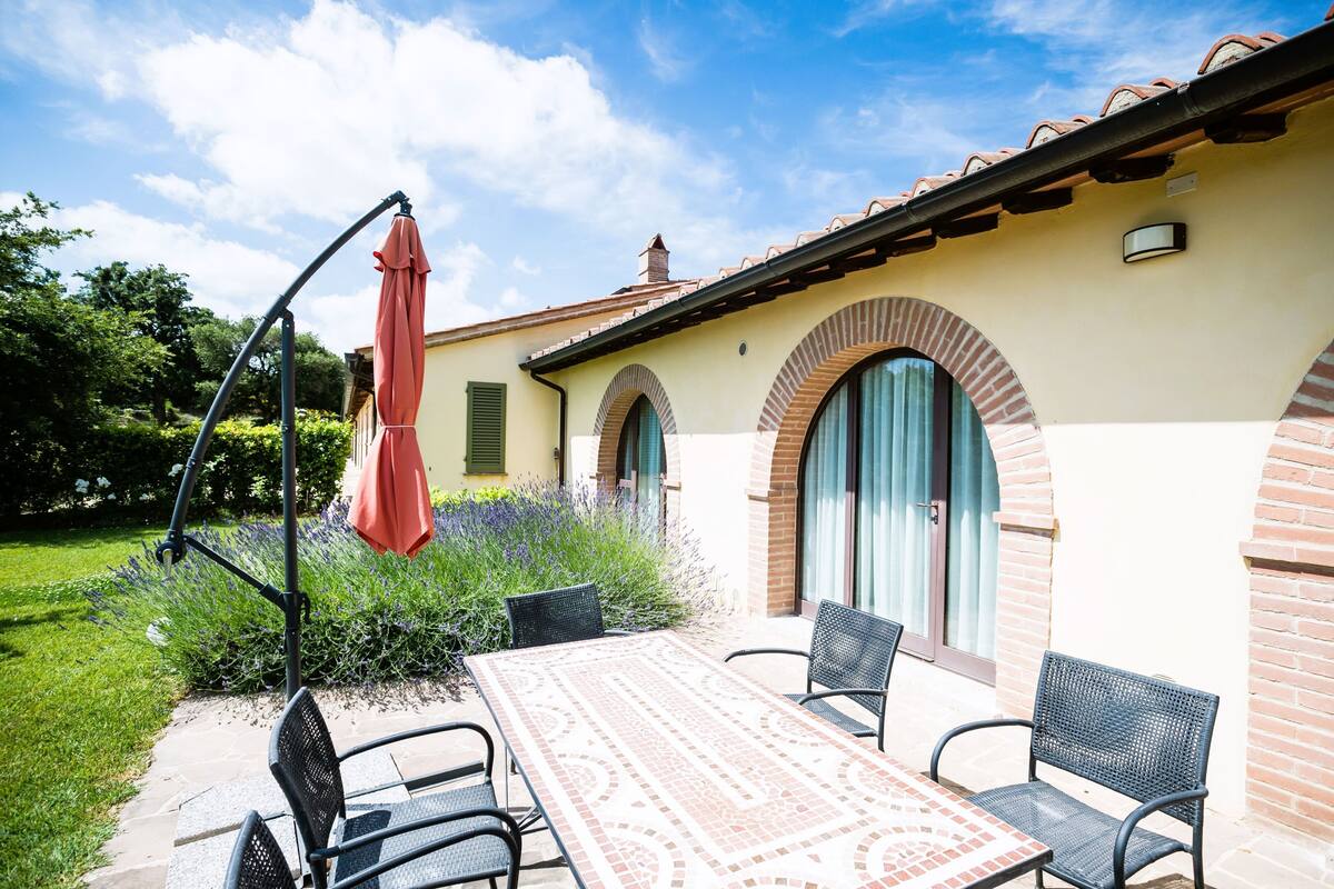 An outdoor patio with a table and chairs, a red umbrella, and a building with arched windows and a tiled roof.