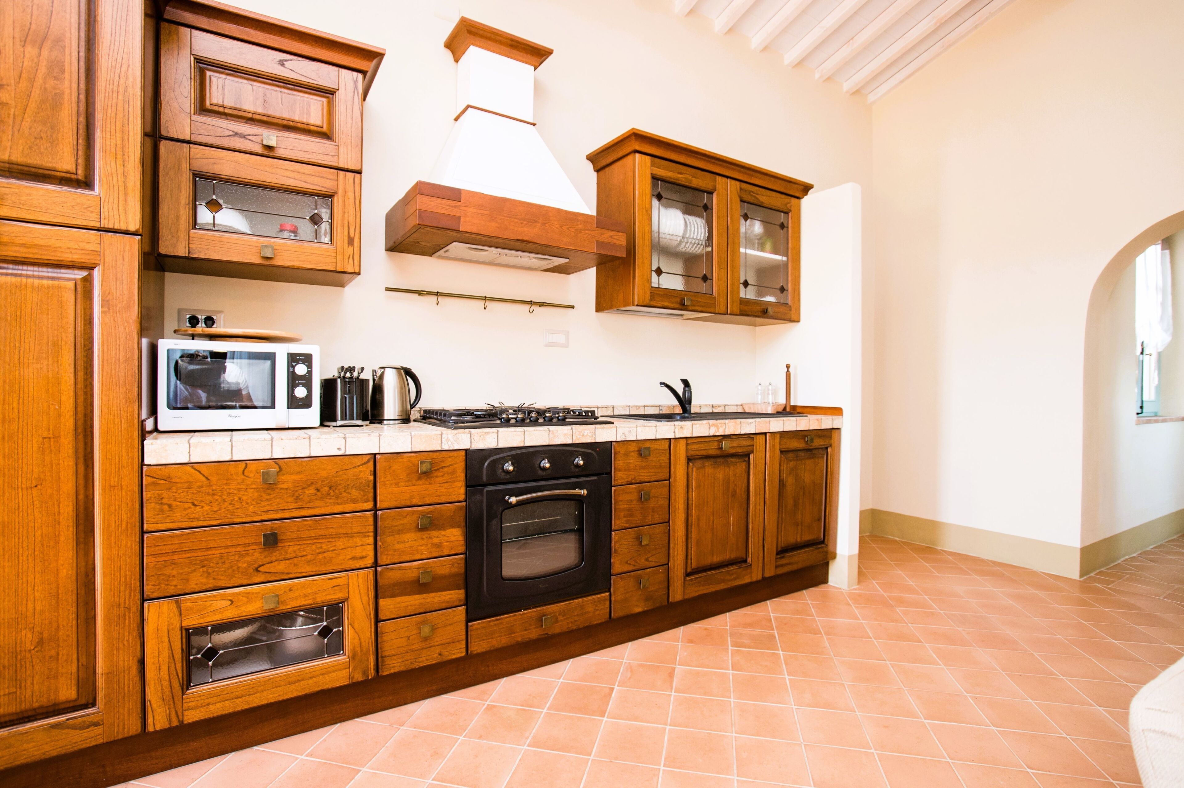 A kitchen with wooden cabinets, a stove, oven, microwave, and a window with curtains.