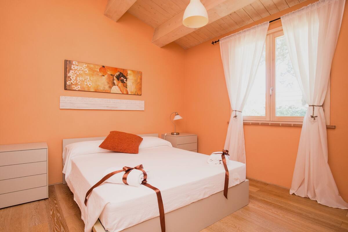 A bedroom with a bed, a nightstand, a lamp, a painting, and a window with curtains.