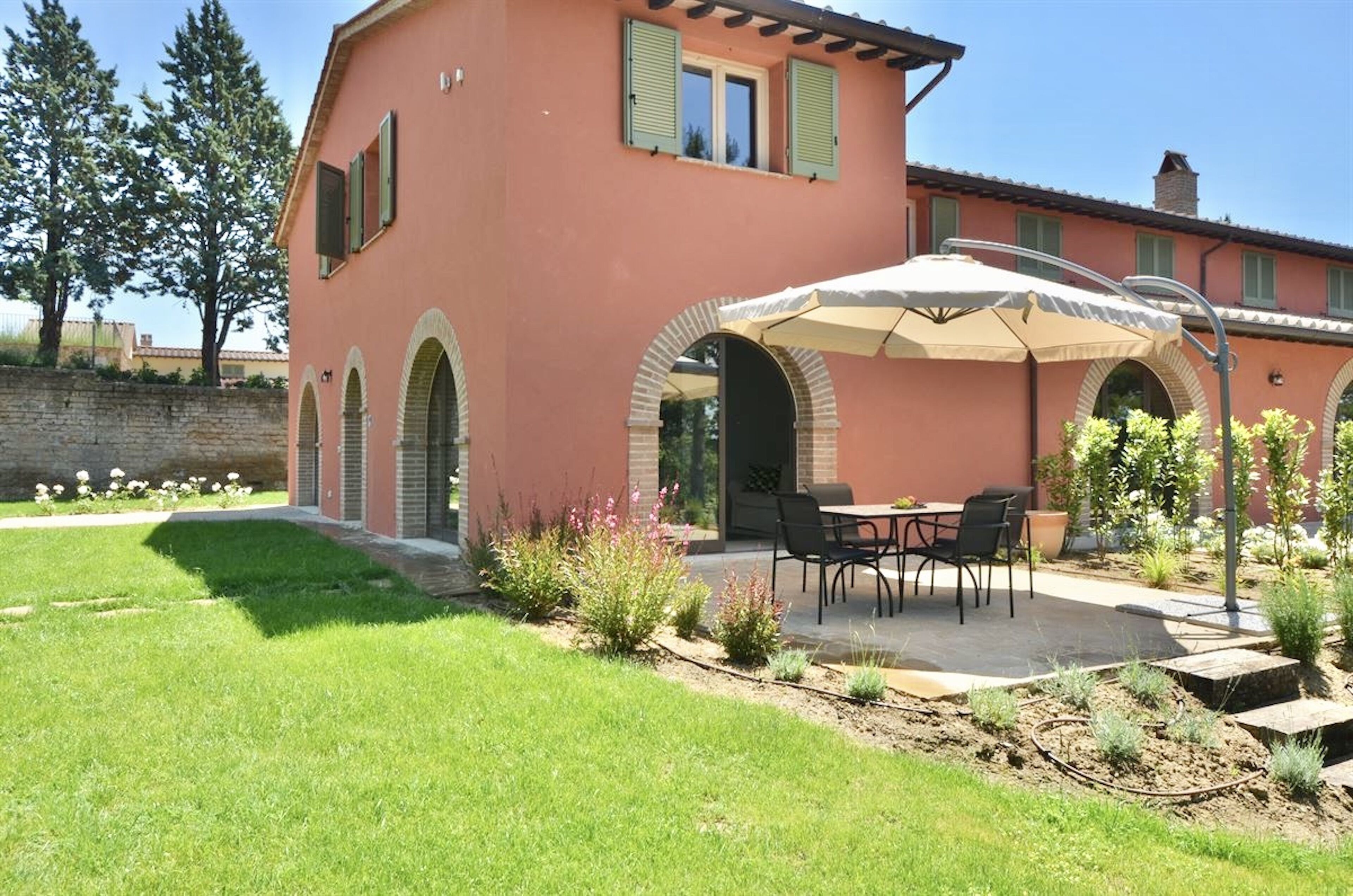 Una casa con un patio arredato da tavolo e sedie, circondata da un giardino con diverse piante e un muro in pietra.