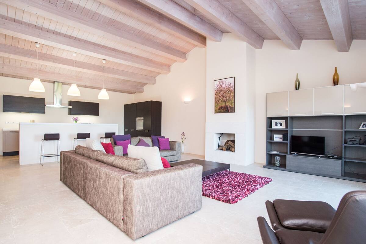 A modern living room with a sofa, a coffee table, and a TV stand. There is a kitchen area with a bar and stools.