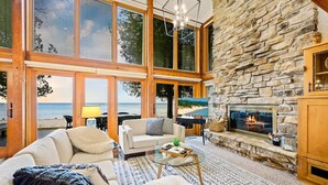 A spacious living room with a stone fireplace, large windows overlooking a beach, and a cozy seating area.