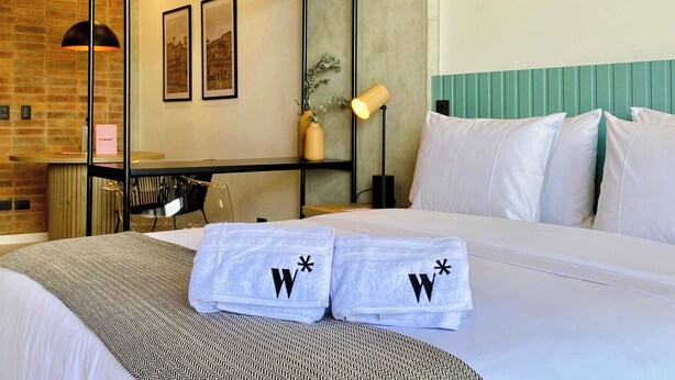 A hotel room with a bed, two towels with a logo, a desk, a chair, a lamp, and framed pictures on the wall. - w* | Marvelous Loft in Polo (Bogotá)