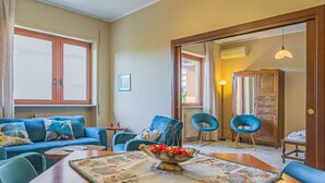 A living room with a blue sofa, two blue armchairs, a wooden dining table with a bowl of fruit, and a large window. - Smile House By Apulia Accommodation (Bari)