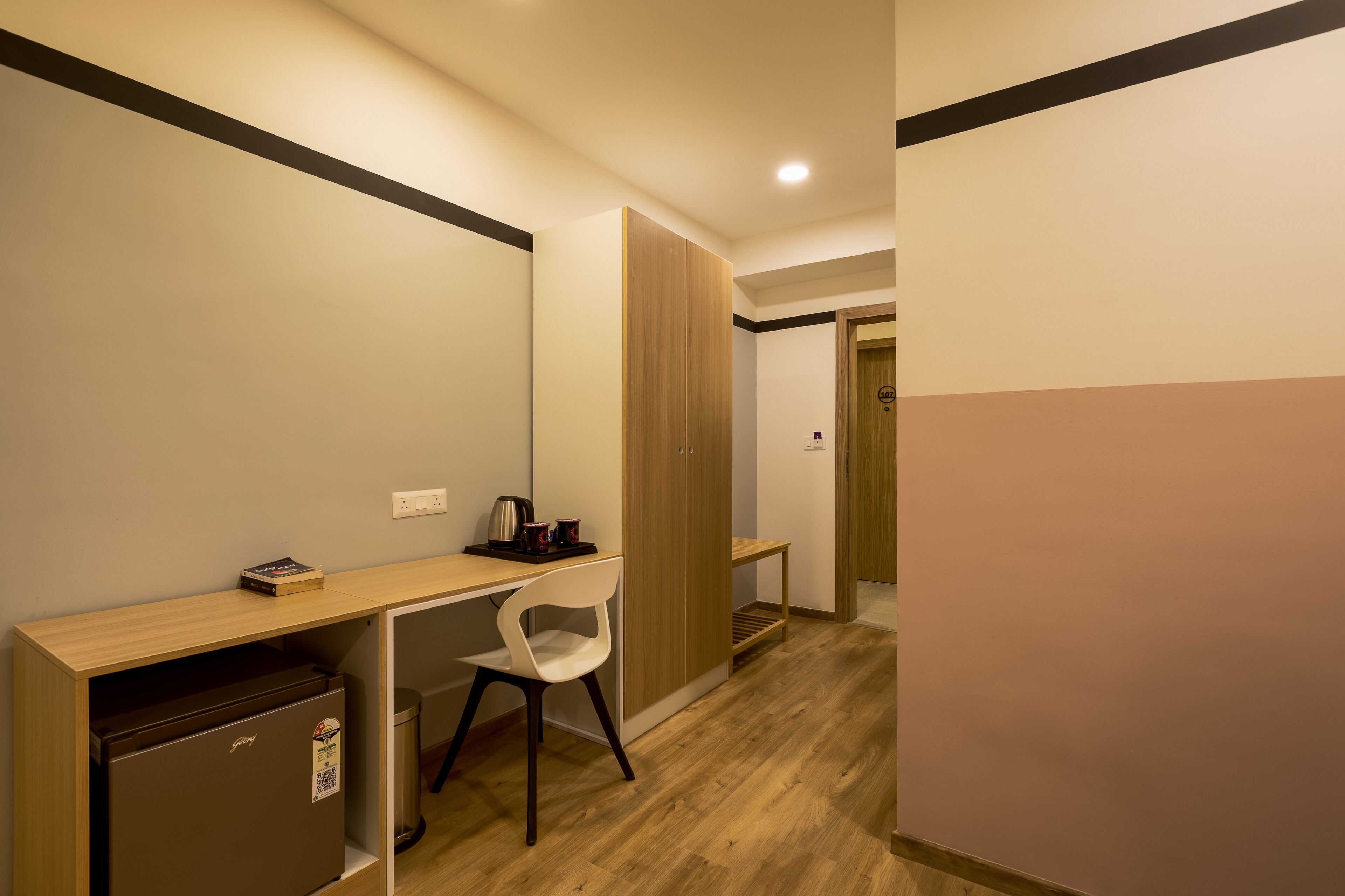 A compact hotel room with a kitchenette, a single white chair, a wooden desk, and a wardrobe.