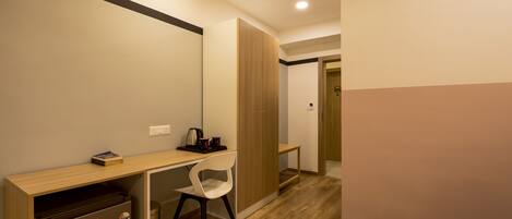 A compact hotel room with a kitchenette, a single white chair, a wooden desk, and a wardrobe.