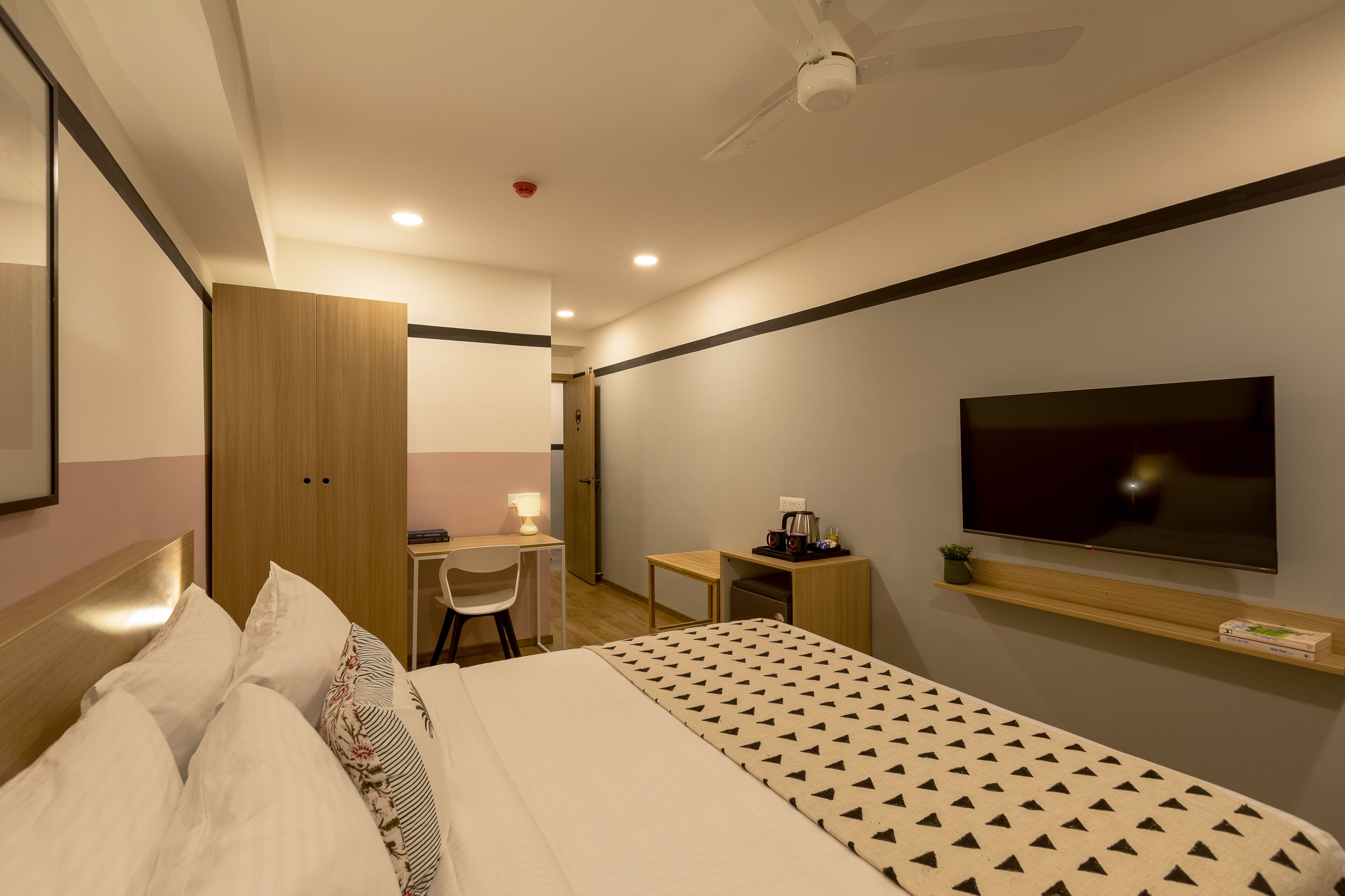 Una habitación de hotel con cama, escritorio, silla, televisión y un pequeño estante.