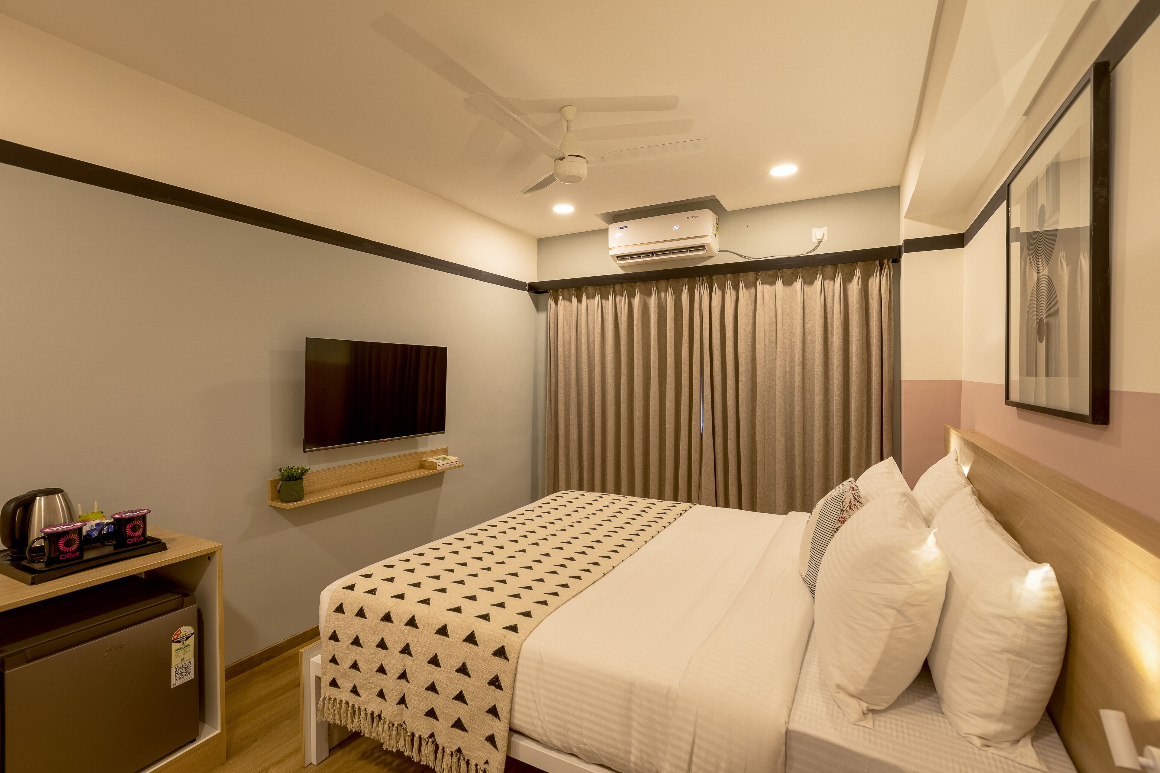 Habitación de hotel con cama, televisión, aire acondicionado, ventilador de techo y escritorio pequeño con tetera y tazas.