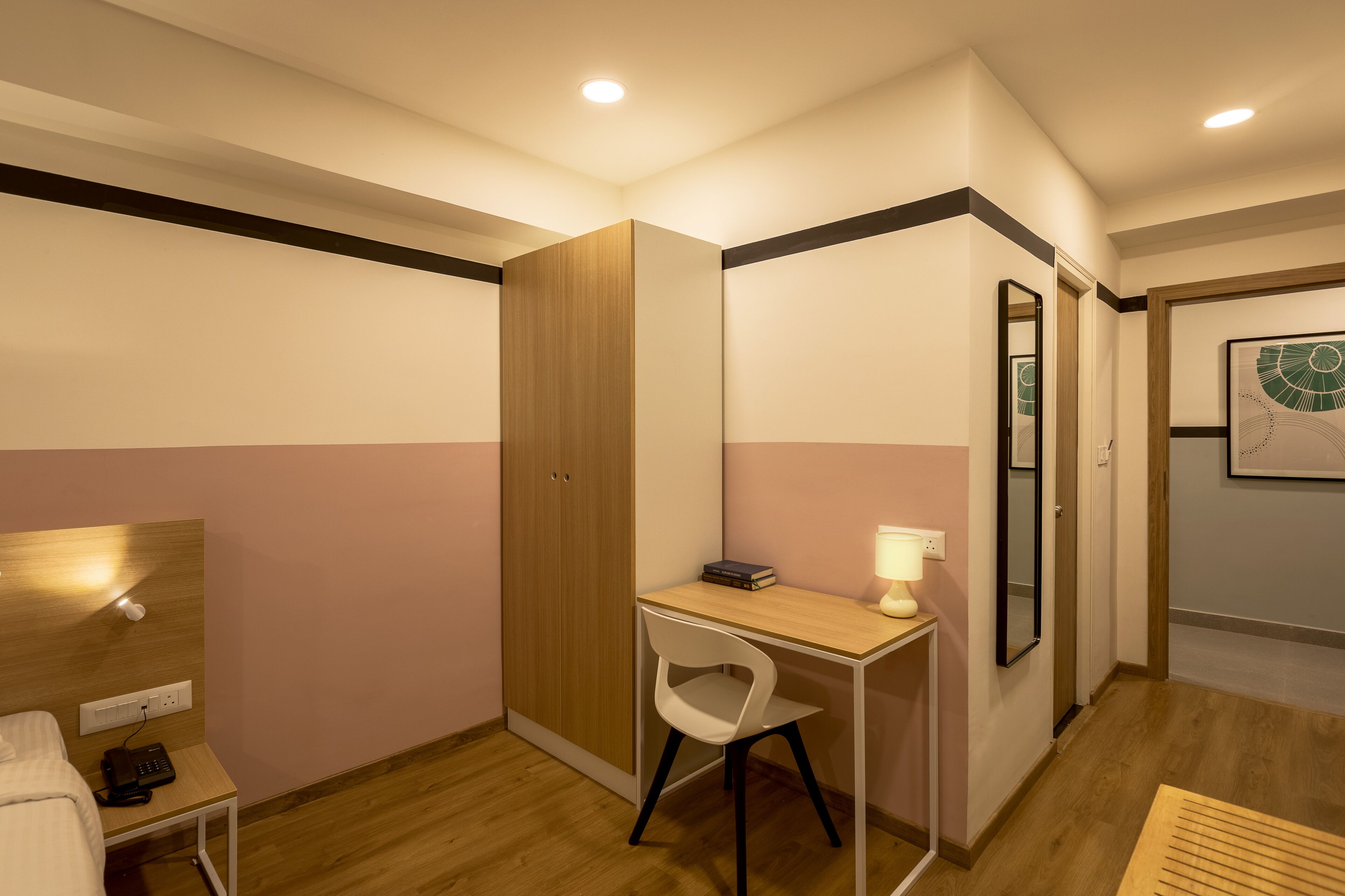 A compact room with a bed, a desk with a chair, a wardrobe, a mirror, and a wall-mounted artwork.